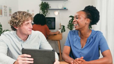 Meslektaşı arka planda bilgisayar üzerinde çalışırken iki profesyonel gülümseyip sohbet ediyor, ortak bir başlangıç atmosferi ve günlük ofis hayatını yakalıyorlar..