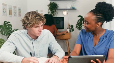 Çeşitli, genç bir ekip, parlak ve modern bir ofiste iş birliği yapıyor. Erkek bir meslektaşım notlar yazarken bayan bir takım arkadaşı arka planda bilgisayar üzerinde çalışan bir meslektaşıyla gülümseyip fikirleri tartışıyor..