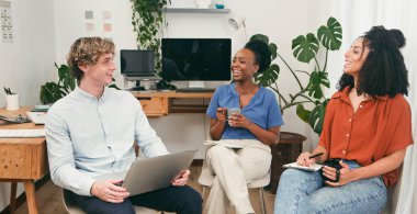 Üç farklı iş arkadaşı parlak, bitki dolu bir ofiste oturuyor. Sohbet eder, gülümser ve dizüstü bilgisayarlar ve defterler etrafında fikirleri paylaşır, takım çalışmasını, yaratıcılığı ve dostane, işbirlikçi bir çalışma ortamını gösterirler..