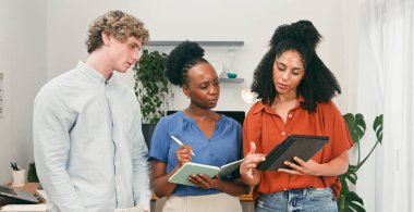 Üç meslektaş parlak ve modern bir ofiste birlikte çalışıyor, fikirleri paylaşıyor ve bir proje planlıyorlar. Takım çalışmasını, öğrenmeyi ve profesyonel işbirliğini yansıtan bir tablet ve defter kullanıyorlar..