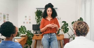 Çiçeklerle dolu parlak bir ofiste, turuncu bluzlu bir kadın konuşmacının iki meslektaşına hitap ettiği sıradan bir iş sunumu. Sahne, yaratıcı bir çalışma ortamında takım çalışması, fikirler ve işbirliği içeriyor..