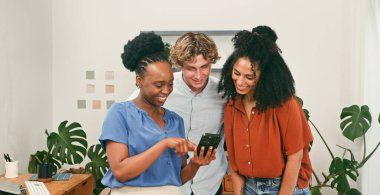 Bir grup arkadaş telefonun etrafında toplanıyor, yeşil bitkiler ve sıcak ışıklarla dolu, parlak ve modern bir çalışma alanında fikirlerini paylaşırken gülümsüyorlar..