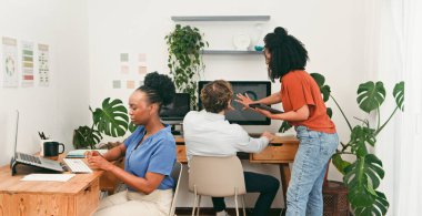 Üç meslektaş, bitkilerle dolu parlak bir ofiste iş birliği yapıyor. Biri bir masada dizüstü bilgisayarla otururken, diğeri bilgisayarda çalışırken, üçüncüsü de tabletle asist yaparak takım çalışmasını ve modern çalışma alanını resmediyor..