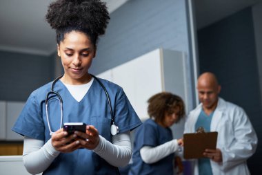 Bir sağlık uzmanı, arka planda hasta belgelerini tartışan meslektaşlarıyla klinik bir ortamda akıllı telefonlarını kontrol ediyor. Takım çalışması ve modern teknoloji tıbbi çevrede verimliliği vurguluyor.