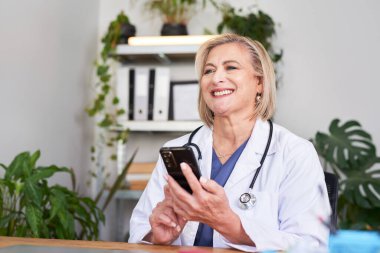 Profesyonel bir kadın doktor, parlak bir klinikteki masada akıllı telefon kullanarak oturur. Sahne, modern sağlık, iletişim ve dost canlısı hasta bakımını kendinden emin ve ulaşılabilir bir ruh haliyle iletiyor..