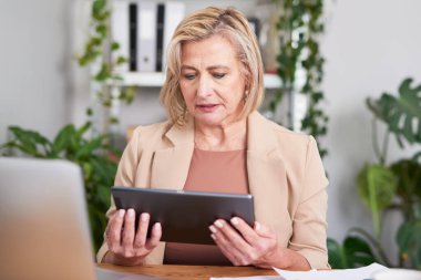 Olgun bir iş kadını, parlak, bitki dolu bir ofiste bir masada oturup, düşünceli bir ifadeyle bir tableti inceliyor. Yakınlardaki dizüstü bilgisayar ve belgeler planlama, analiz ve profesyonel çalışmaların devam ettiğini gösteriyor..