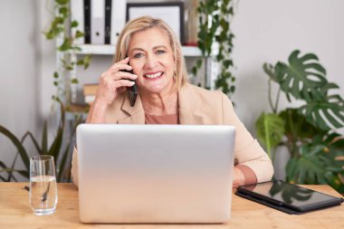 Gülümseyen bir iş kadını, parlak bir ofisin ahşap masasında oturmuş, telefonda bir dizüstü bilgisayar ve tabletin yanında konuşuyor. Yeşillik bitkiler ve organize raflar sakin ve üretken bir çalışma ortamı yaratır.