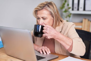 Odaklanmış profesyonel bir kadın, yoğun bir iş gününden kahve molası vererek, modern bir ofiste dizüstü bilgisayarının başında çalışıyor. Sahne üretkenlik, konsantrasyon ve sakin bir iş ortamı sunuyor..