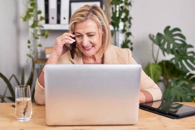 Profesyonel bir kadın parlak bir ofiste ahşap bir masada oturur, cep telefonuyla konuşur ve gümüş bir dizüstü bilgisayara odaklanır. Bitkiler ve ofis malzemeleri sakin ve üretken bir atmosfer yaratır..