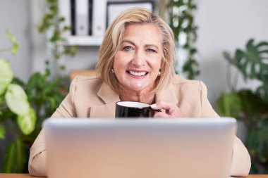 Bej ceketli gülümseyen olgun bir kadın elinde kupa olan bir dizüstü bilgisayarla bir masada oturuyor. Parlak ofis bitkileri iş için elverişli ve davetkar bir ortam yaratıyor..