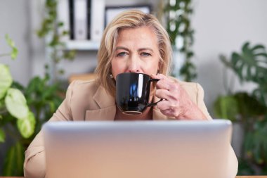 Bej ceketli profesyonel bir kadın laptopuna odaklanmış siyah bir fincandan kahve içiyor. Bereketli bitkilerle dolu parlak ofis ortamı sakin ve üretken bir atmosfer yaratır..