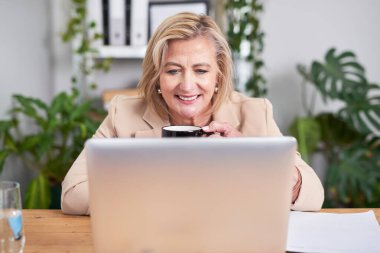 Olgun profesyonel bir kadın ahşap bir masada oturur ve bir fincan kahvenin tadını çıkarırken dizüstü bilgisayarına bakar. Yeşillik bitkiler ve düzenli bir çalışma alanı sakin ve verimli bir ofis ortamı yaratır..