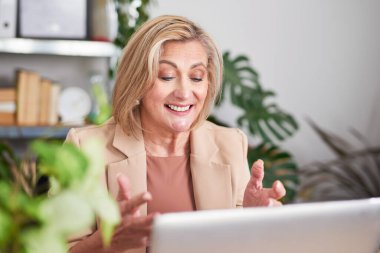 Bej ceketli, gülümseyen profesyonel bir kadın, üretkenliği, iletişimi ve pozitif iş enerjisini aktaran, bitkilerle dolu parlak bir ofiste dizüstü bilgisayarda çalışır..