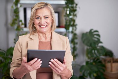 Profesyonel, gülümseyen bir kadın, parlak ve modern bir ofiste tablet tutuyor. Yeşillik bitkilerle çevrelenmiş olarak, kendine güven, verimlilik ve yaklaşımı iletir - iş, liderlik ve uzak çalışma temaları için mükemmeldir..