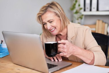 Modern bir ofiste masa başında oturan profesyonel bir kadın kahvesini yudumlarken dizüstü bilgisayarına odaklanıyor. Sahne üretkenlik, konsantrasyon ve sakin bir iş ortamı sunuyor..