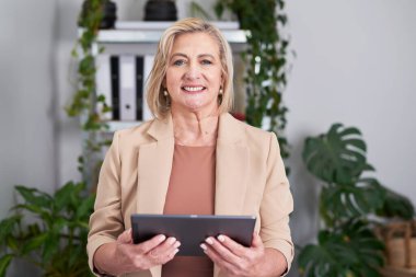 Profesyonel, gülümseyen, bej ceketli yaşlı bir kadın yemyeşil bitkilerle dolu parlak bir ofiste tablet tutuyor. Bu stok fotoğrafı liderlik, profesyonellik ve yaklaşılabilir bir kurumsal titreşim taşır..