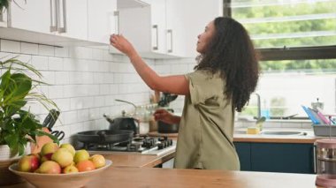 Kıvırcık saçlı genç bir kadın, parlak, modern bir mutfakta kahve makinesini kullanarak sabah kahvesini yapmaya hazırlanan kabineden bir kupa alır..