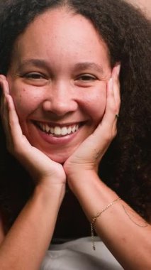 Doğal kıvırcık saçlı güzel bir siyah kadın kameraya gerçekten gülümsüyor, elleri nazikçe yüzünü kavrıyor. Mutluluk, pozitiflik ve memnuniyet taşıyor. Bu yakın portrede neşe saçıyor..