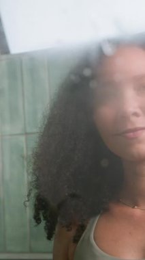 Odaklanmış, doğal saçlı, sarı eldivenli, sisli cam bir yüzeyi silikonla temizleyen siyah bir kadın. Gülümsemesi memnuniyeti yansıtıyor, cam tamamen berraklaşıyor, evde bakım ve verimlilik gösteriyor..