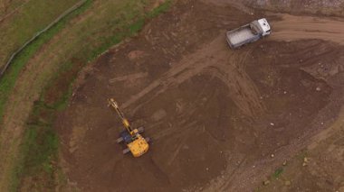 Toprak kazan sarı kazıcının yukarıdan aşağı hava görüntüsü ve toprak hareket halindeki bir inşaat alanındaki beyaz bir çöp kamyonuna çakıl yükleme.