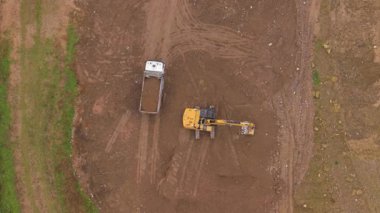 Sarı kazıcının yukarıdan aşağıya havadan görüntüsü. İnşaat alanında toprağı hareket ettiriyor. Toprakla dolu beyaz bir çöp kamyonu bölgeden uzaklaşıyor.