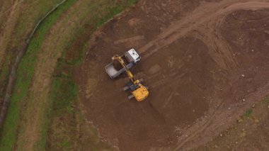 Sarı kazıcının yukarıdan aşağıya hava görüntüsü toprağı kazıyor ve beyaz bir çöp kamyonuna yüklüyor. Toprak hareketinde çalışan ağır makineler