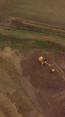Yeşil bir tarlanın yanındaki bir toprak parçasının üzerine park edilmiş büyük sarı bir kazıcının hava görüntüsü. Toprağı hareket ettirmeye ve kazmaya hazırlanıyor.