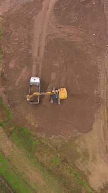 Ağır makinelerin havadan aşağı görüntüsü iş başında. Sarı bir kazıcı toprağı kazar ve dikkatlice büyük bir çöp kamyonunun arkasına koyar.