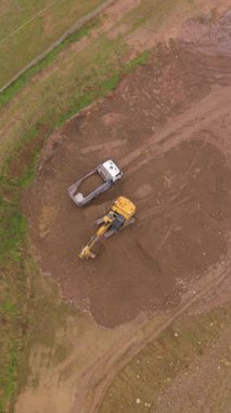 Sarı kazıcı toprak kazıyor ve bir inşaat alanındaki çöp kamyonuna toprak yüklüyor. Ağır makinelerin çalışan tepeden tırnağa görüntüsü