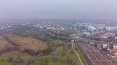 Büyük bir demiryolu istasyonu ve raylarının yanında büyük bir halk parkı bulunan şehir manzarası sislerin arasından görülebiliyor.
