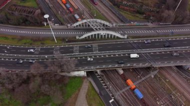 Kompleks bir ulaşım merkezinin yüksek açılı insansız hava aracı perspektifi, çok şeritli bir otoyolda arabaları ve bitişik tren raylarında yük trenlerini gösteriyor