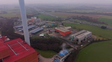 Yüksek açılı bir modern sanayi tesisi manzarası, uzun bacası olan, buhar veya duman çıkaran, kırsal bir arazide otoban olan.