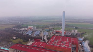 Kırmızı çatıları ve atmosfere buhar salan uzun beyaz bacası olan endüstri kompleksi. Çevreleyen yeşil alanlar ve arka planda yoğun bir otoyol