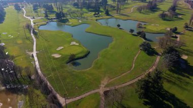 Güzel bir golf sahasında uçan yüksek açılı drone perspektifi. Bereketli yeşil çimenler, kum sığınakları, ağaçlar ve büyük bir göl manzaralı bir manzara yaratır.
