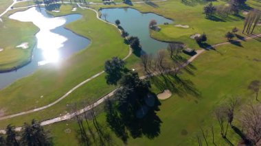 Güneşli bir günde yeşil bir golf sahasında uçan bir hava aracı. Manzara, yemyeşil yollar, ışıl ışıl göller ve kum sığınaklarını kapsıyor.