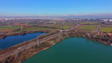 Sersemletici hava manzarası iki yapay gölün bir otoyol ve demiryolu ile bölündüğünü gösteriyor. Milano şehrinin silueti ve arka planında Alpler var.