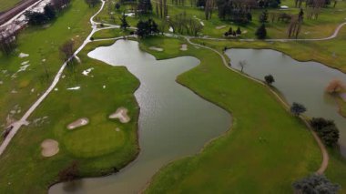 Güzel yeşil bir golf sahasının geniş bir hava görüntüsü. Manikürlü çim yolları, kum sığınakları ve yansıtıcı göller yer almaktadır.