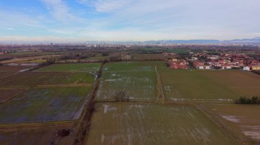 Sel basmış tarlaları ve ekili arazileri olan geniş bir kırsal alan manzarası. Arka planda, uzak bir şehrin silueti görünür.