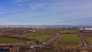 İtalya kırsalında seyahat eden bir banliyö treninin hava perspektifi, arka planda Milan şehri ve Alpler ile bir köprünün altından geçiyor.