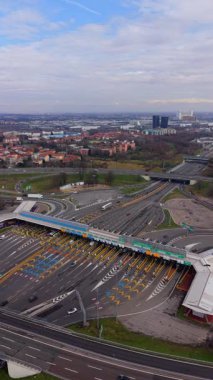Yüksek açılı insansız hava aracı görüntüleri büyük bir otoyol gişesinden geçen araba ve kamyonları gösteriyor. Arka planda modern yol altyapısı ve şehir manzarası