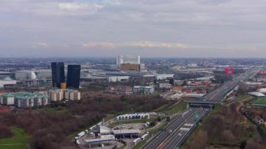 Trafiği akan büyük bir otoyolun geniş hava manzarası. Şehir modern binalar, bir benzin istasyonu ve bir park ile aydınlanır.