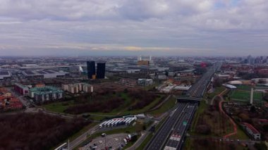 Milano, İtalya 'ya giden arabalar ve kamyonlarla dolu yoğun bir otoyolu gösteren hava aracı görüntüsü. Şehir manzarası bulutlu bir günde modern binaları ve yeşil alanları içerir.