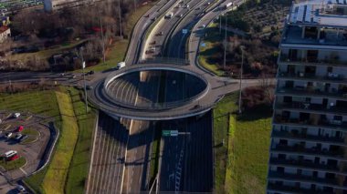 Modern dairesel bir kavşağın dinamik hava perspektifi. Araçlar çok şeritli otoyolda ve şehir binaları yakınlarındaki kavşakta ilerliyorlar.
