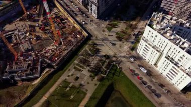 Yüksek açılı insansız hava aracı modern bir şehrin üzerinde uçuyor. Manzara, caddelerde giden konut binaları ve arabaların yanındaki büyük bir inşaat alanını yakalıyor.