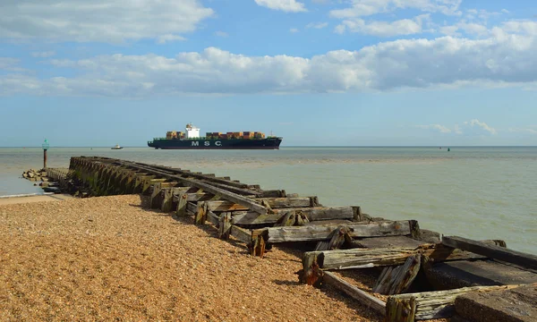 Un pequeño barco de contenedores que entra en el puerto de Felixstowe siendo guiado por remolcador, viejo rompeolas en primer plano . — Foto de Stock