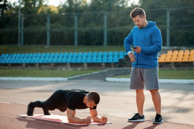 Adam dışarıda kişisel fitness eğitmeniyle kalas egzersizi yapıyor.
