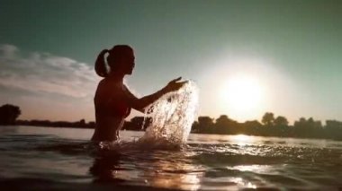 Kırmızı bikinili mutlu kız su sıçratır veya suda oynar ve gün batımında eğlenir.