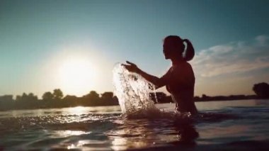 Kırmızı bikinili mutlu kız su sıçratır veya suda oynar ve gün batımında eğlenir.