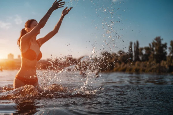 Kırmızı bikinili mutlu bir kadın su sıçratıyor ya da suda oynuyor ve gün batımında eğleniyor.