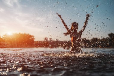 Kırmızı bikinili mutlu bir kadın su sıçratıyor ya da suda oynuyor ve gün batımında eğleniyor.